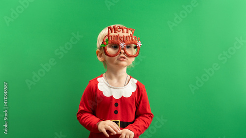  Happy Toddler girl in red Christmas costume and glasses smiling on green background COPYSPACE