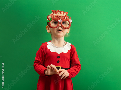  Happy Toddler girl in red Christmas costume and glasses smiling on green background COPYSPACE