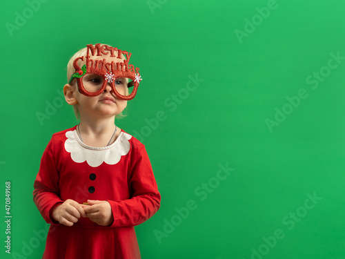  Happy Toddler girl in red Christmas costume and glasses smiling on green background COPYSPACE