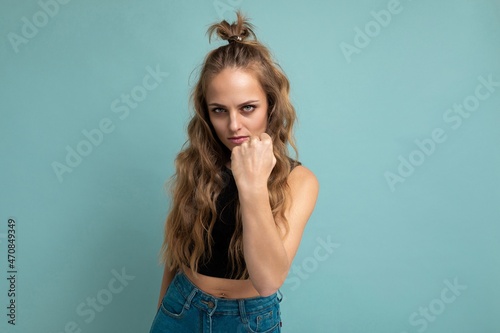 Portrait photo of young beautiful pretty self-confident angry blonde woman with sincere emotions wearing stylish black top isolated over blue background with free space and showing fist at the camera © Ivan Traimak