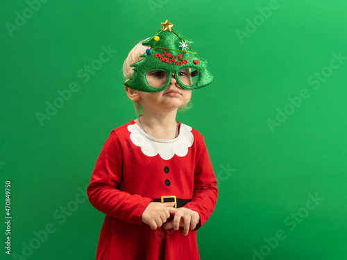  Happy Toddler girl in red Christmas costume and green tree glasses smiling on green background COPYSPACE
