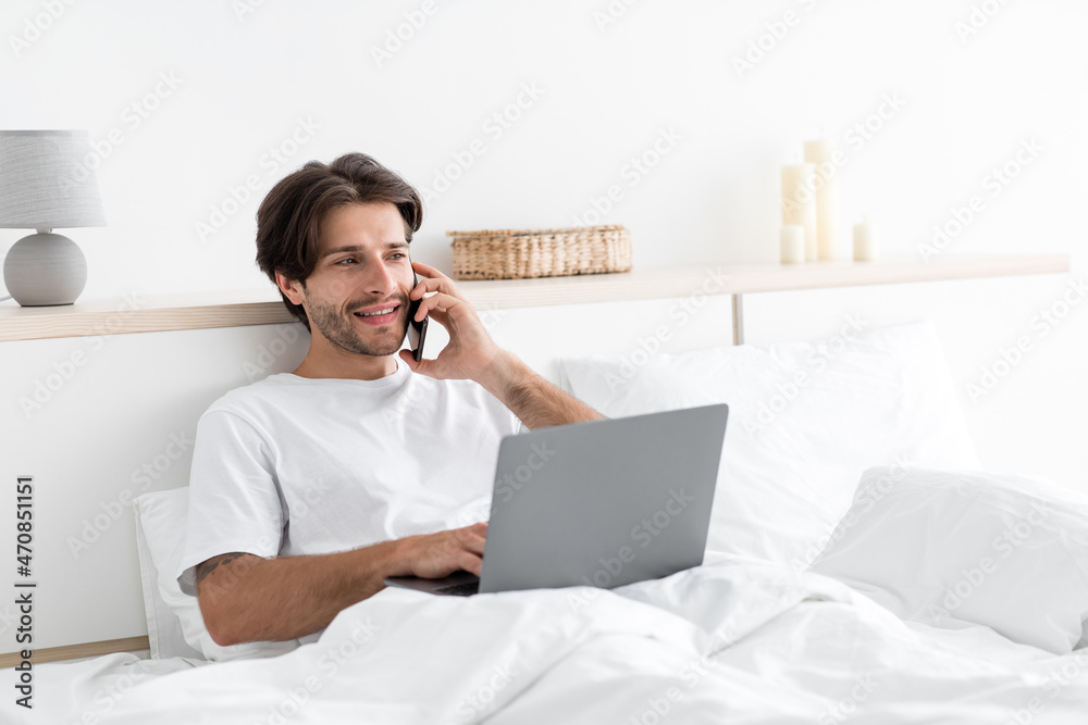 Smiling handsome millennial caucasian guy works on laptop, speaks by phone with client Smiling handsome millennial caucasian guy works on laptop, speaks by phone with client