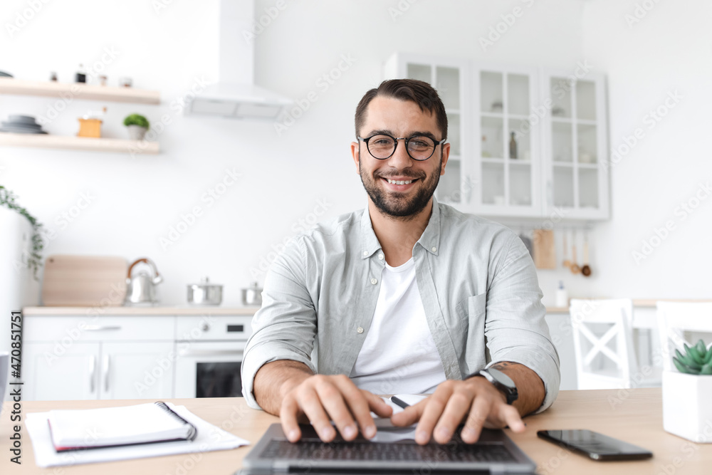 Smiling mature caucasian man in glasses working at computer on kitchen interior