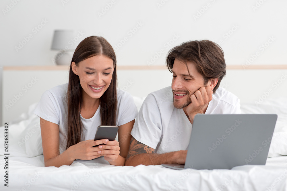 Happy young european family lie on bed, with laptop and smartphone chatting, watch video or shopping Happy young european family lie on bed, with laptop and smartphone chatting, watch video or shopping