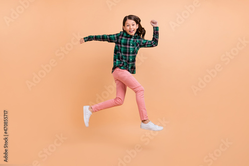 Photo of sweet funky school girl dressed checkered clothes smiling running fast jumping high isolated beige color background
