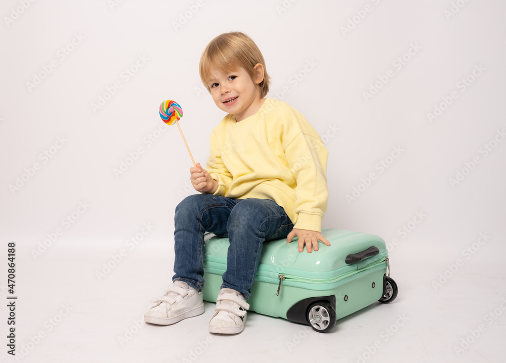 Happy little boy holding lollipop sits in a suitcase on a white background with space for text