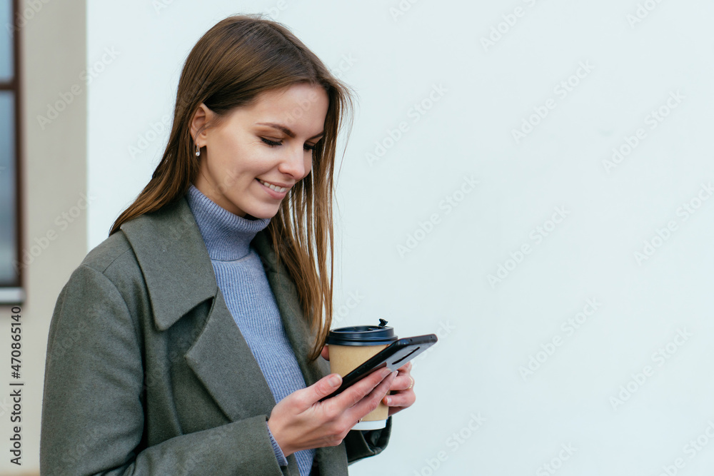 Beautiful smiling girl with cofe cup in street, holding smartphone in hands and looking to device Beautiful smiling girl with cofe cup in street, holding smartphone in hands and looking to device