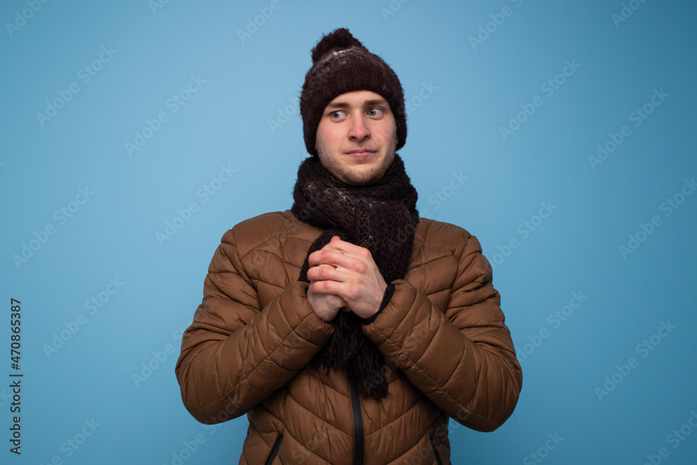 Surprised young man in brown mittens and a hat, light brown jacket on a blue background, studio portrait Surprised young man in brown mittens and a hat, light brown jacket on a blue background, studio portrait