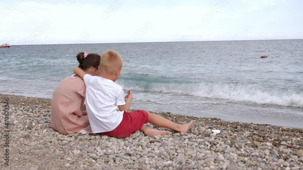 A cute little boy is sitting on the beach next to a girl, he hugs her shoulders and together they throw stones into the sea. A couple of children are relaxing on the beach at sunset. A cute little boy is sitting on the beach next to a girl, he hugs her shoulders and together they throw stones into the sea. A couple of children are relaxing on the beach at sunset.