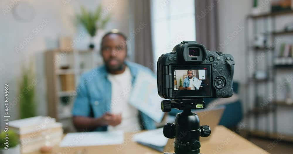 Focus on camera on which likable confident skilful young bearded african american in wireless headset explaining diagram for his internet audinece from home office