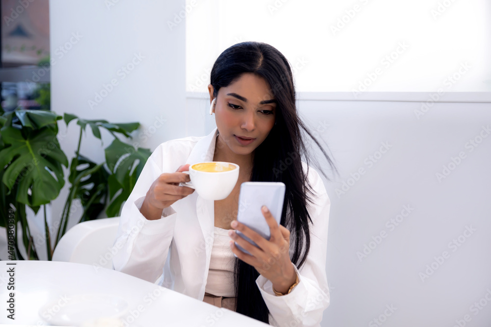 Young Indian ethnicity woman with long dark hair using smart phone and drinking coffee, lifestyle Young Indian ethnicity woman with long dark hair using smart phone and drinking coffee, lifestyle