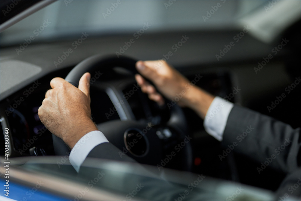 Naklejka premium Close up of male client in stylish suit sitting on driver's seat with hands on steering wheel while testing car at showroom. Concept of people, purchase and selling.
