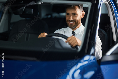 Smiling bearded man in formal clothes sitting inside modern car and examining leather interior. Happy male client choosing new vehicle at showroom. © serhiibobyk