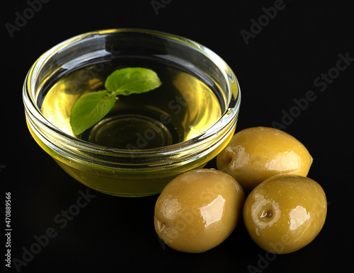Olive oil in a glass bowl with basil leaf and large green olives on a black background. Top view