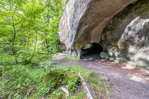 Fototapeta Naklejka Na Ścianę i Meble -  Mullerthal Trail with a huge rock formation with a tunnel, uneven and weathered surface, trees and green wild vegetation, summer day on one of the hiking trails in Luxembourg