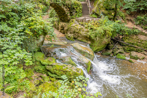 Black Ernz river among wild vegetation  Schiessent  mpel waterfall seen from a higher perspective  crystal clear water flowing between rocks  staircase in the background  Mullerthal Trail  Luxembourg