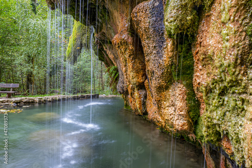 Calcareous crystalline water falling from a moss covered rock formations into a basin in the Kallektuffquell waterfall, trees in the background, Mullerthal Trail, Luxembourg. Long exposition image