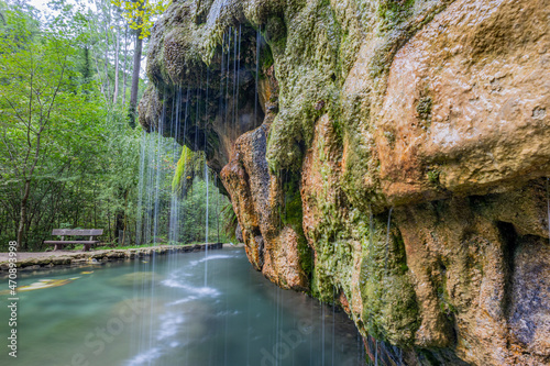 Crystal clear calcareous water flowing between moss covered rock formations in a basin, Kallektuffquell waterfall, bench and trees in the background, Mullerthal Trail, Luxembourg. Long exposition photo