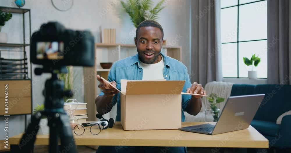 Attractive smiling happy bearded african american blogger filming new video on videocamera for his vlog when unpacking carton box with purchase at home
