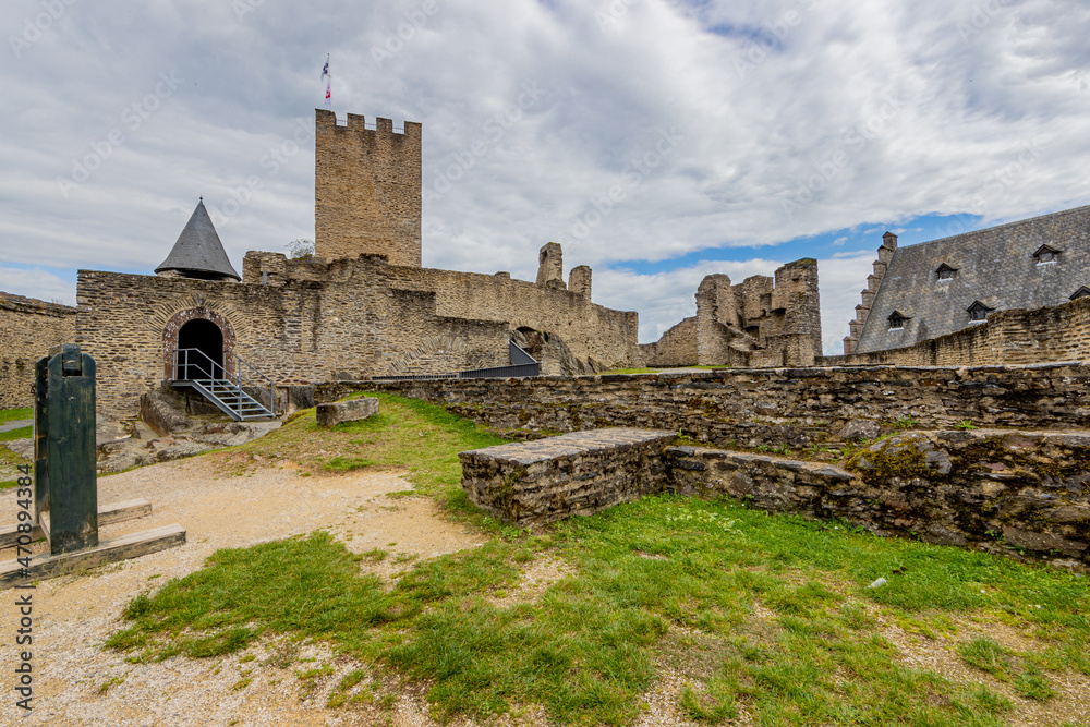 Open-air ruin at medieval Bourscheid castle, courtyards, green grass, ruined stone walls, a tower, cellar and stairwell, cloudy day with a blue sky and gray clouds, Luxembourg