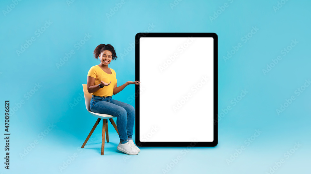 Black female sitting on chair, pointing at giant tablet pc with white screen, offering space for app or website, mockup Black female sitting on chair, pointing at giant tablet pc with white screen, offering space for app or website, mockup