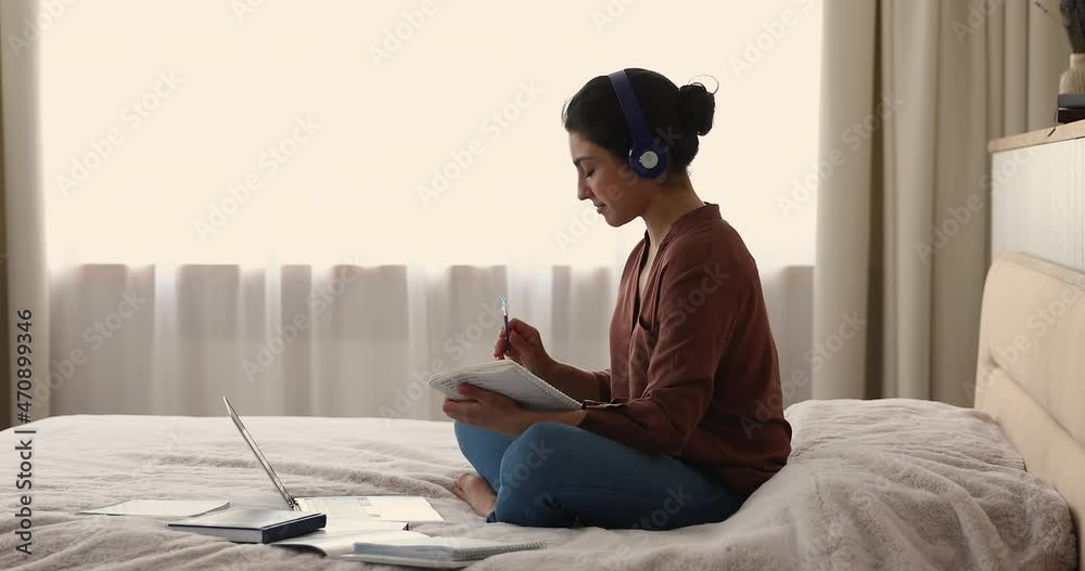Smart young Indian female student in wireless headphones sit on large bed cross legged study from home before laptop pc. Millennial lady trainee hold dialogue with mentor in conference app take notes