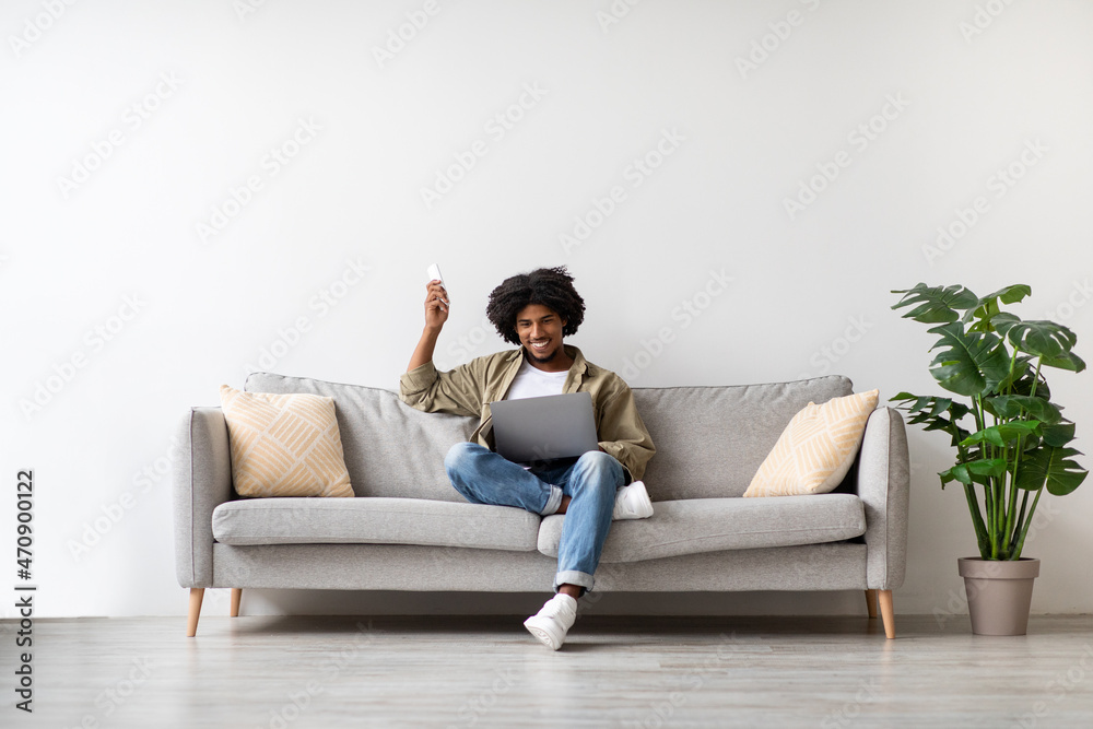 Obraz premium Young Black Man Sitting On Couch With Laptop And Using Remote Controller