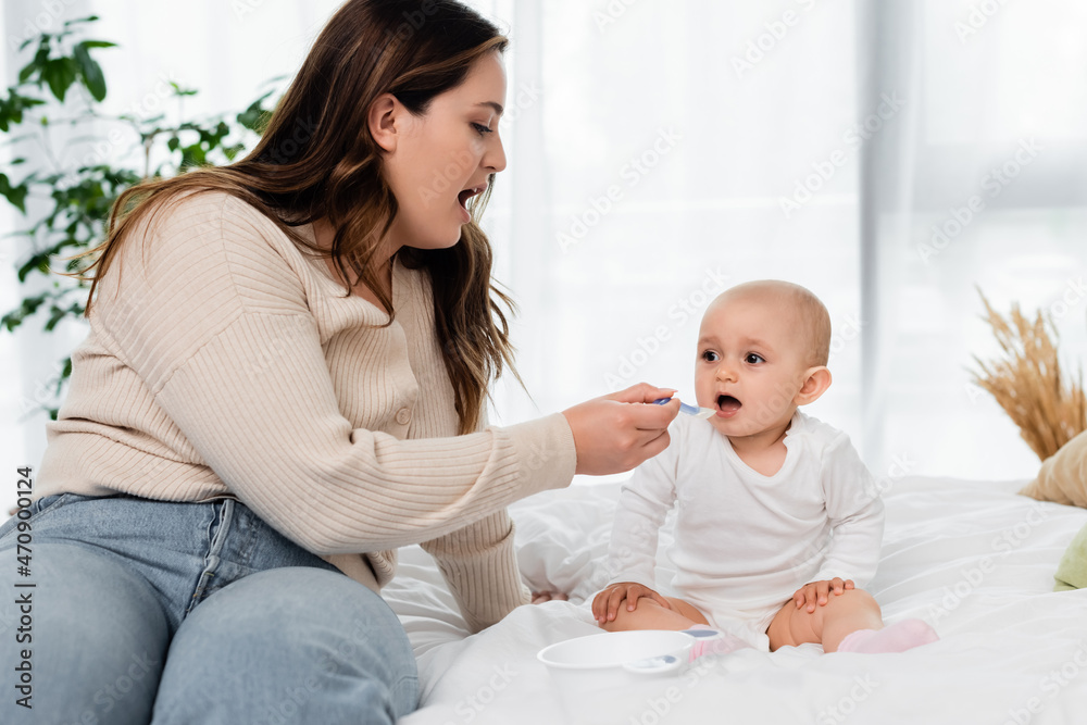 Plus size mother open mouth while feeding baby on bed.