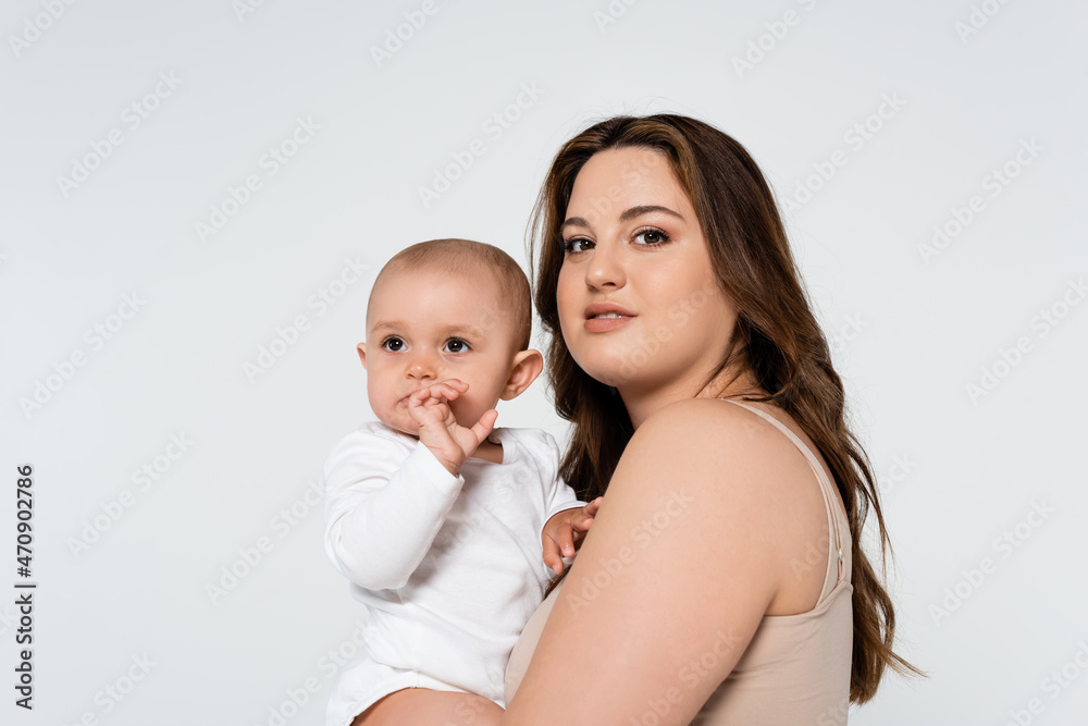 Pretty body positive woman hugging baby girl and looking at camera isolated on grey. Pretty body positive woman hugging baby girl and looking at camera isolated on grey.