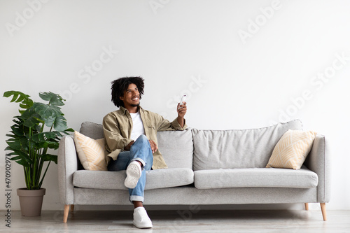 Happy Black Guy Sitting On Couch, Operating Air Conditioner With Remote Controller