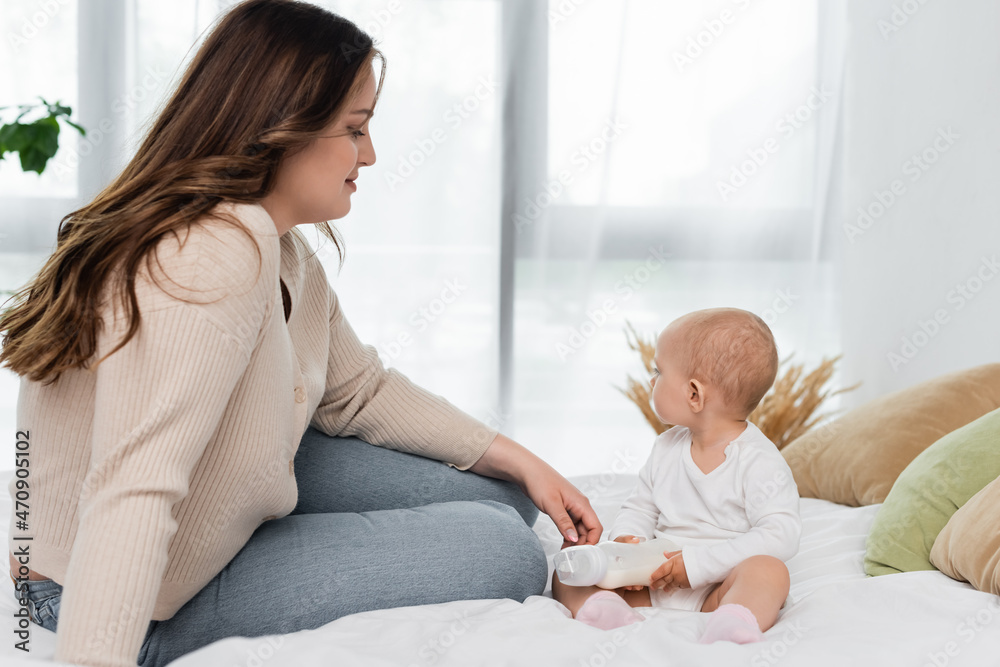Fototapeta premium Pretty pus size woman sitting near baby daughter with bottle of milk on bed.