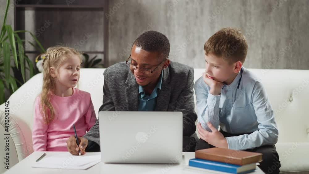 Positive African-American English teacher conducts private lesson to junior schoolchildren writing letters near laptop in living room slow motion Positive African-American English teacher conducts private lesson to junior schoolchildren writing letters near laptop in living room slow motion