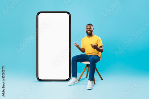 Black man sitting on chair next to huge smarthone with blank white screen, advertising new mobile application, mockup