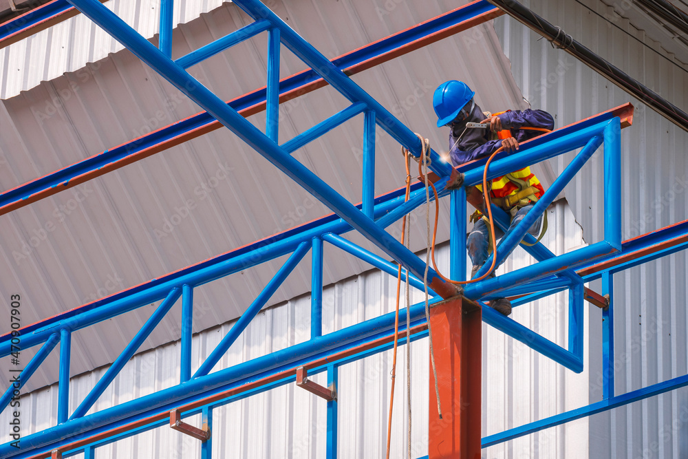 Low angle view of construction worker with safety equipment is welding metal roof warehouse building structure in construction site