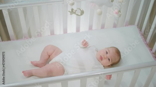 Beautiful baby girl relaxing on her baby cot alone looking up at the toys cot and playing with hands very cute. 4k