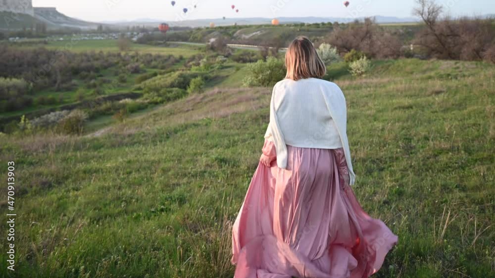 Red haired thin beautiful young girl in a pink long dress fluttering in the wind and a white jacket falling from her shoulders runs down the slope to the colorful balloons