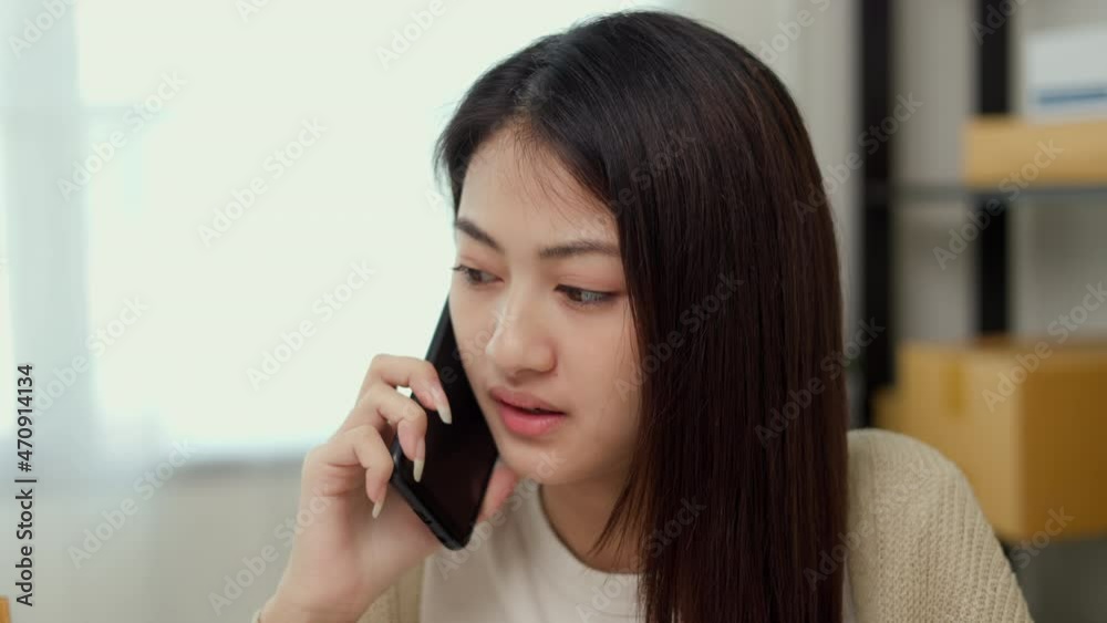 Young asian business woman using mobile phone call receiving purchase order and check product on stock. Sit near the package shelf on background. Ecommerce business and shopping online concept. Young asian business woman using mobile phone call receiving purchase order and check product on stock. Sit near the package shelf on background. Ecommerce business and shopping online concept.