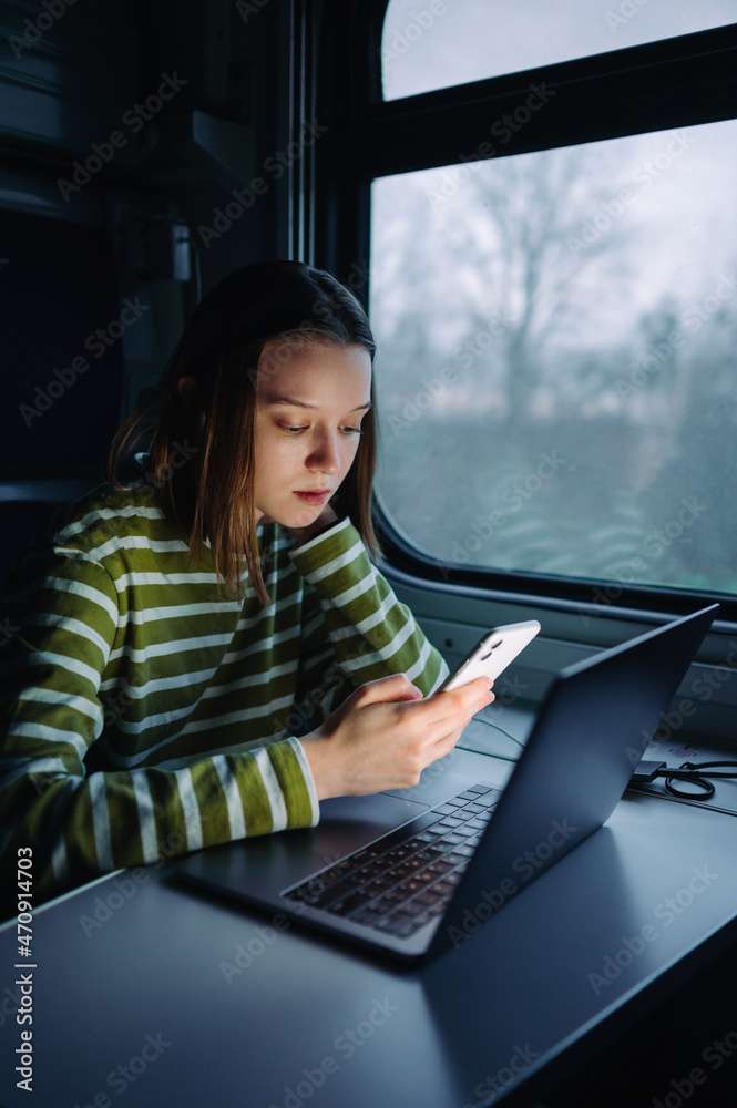 Beer woman sitting in a train with a laptop and uses a smartphone with a serious face. Vertical