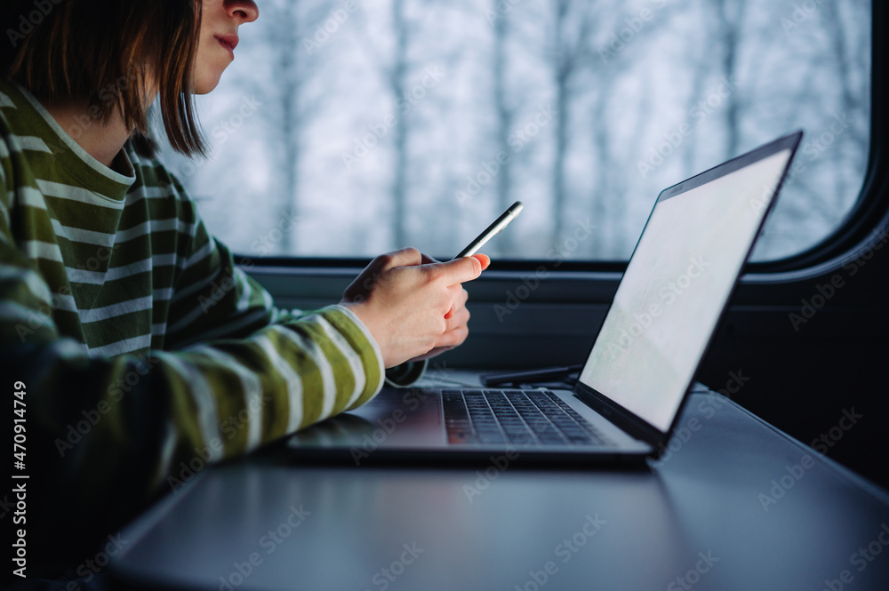 A woman sits at a table in the evening in a train and uses a smartphone and works on a laptop. Working on a laptop while traveling.