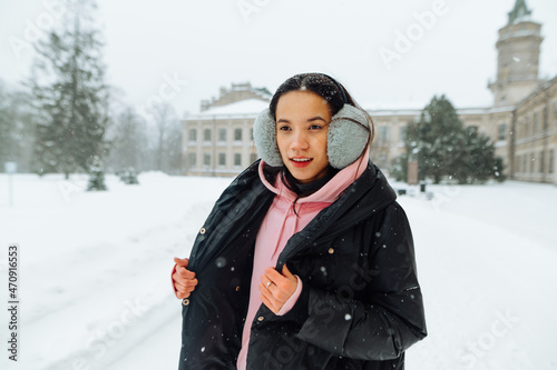 Smiling hispanic woman in warm casual clothes standing in the park near beautiful historic architecture in winter and smiling looking away.