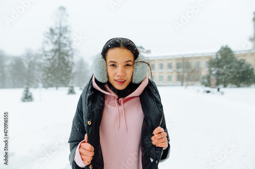 Cute positive hispanic woman standing in a winter park during a snowfall and posing for the camera with a smile on her face.