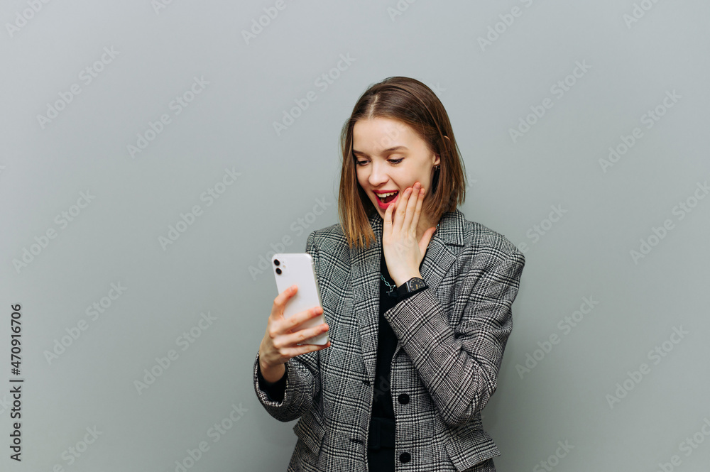 Joyful woman with a shocked face holds a smartphone in her hands and looks at the screen with delight on a gray background.