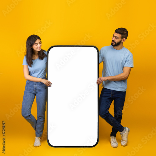 Cool mobile app. Millennial arab spouses standing near giant smartphone with mockup, promoting app or website