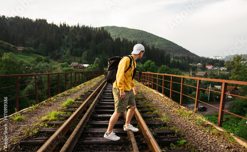 Young male tourist stands on a railway viaduct in the mountains against the backdrop of a beautiful landscape and looks to the side, wearing an orange sweatshirt.