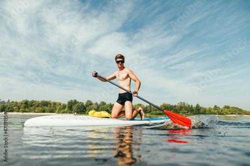 Muscular athletic man in sunglasses actively rowing with an oar sitting on a sup board, swims on a pond. Summer fun on the water.