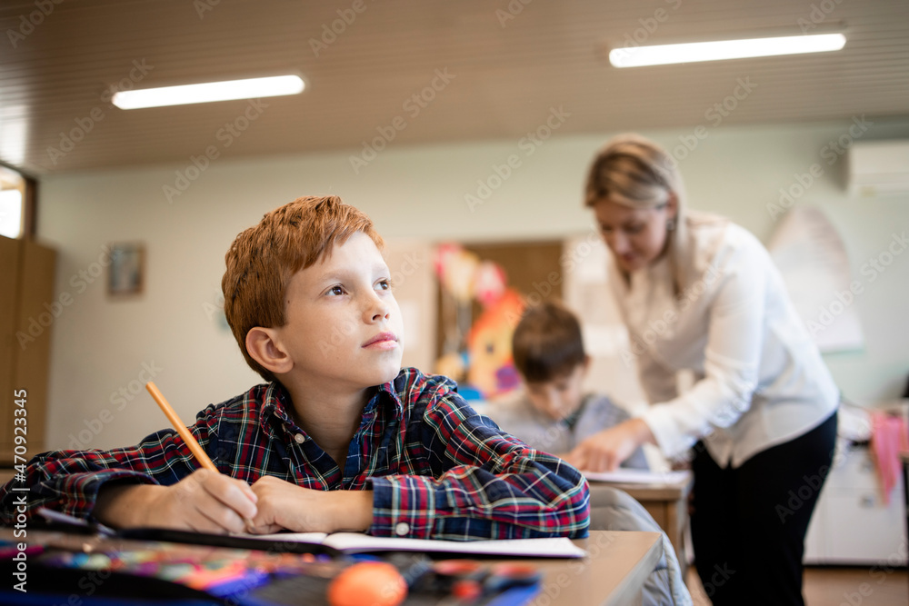Portrait of smiling caucasian schoolboy thinking about correct answer in classroom and teacher in background.