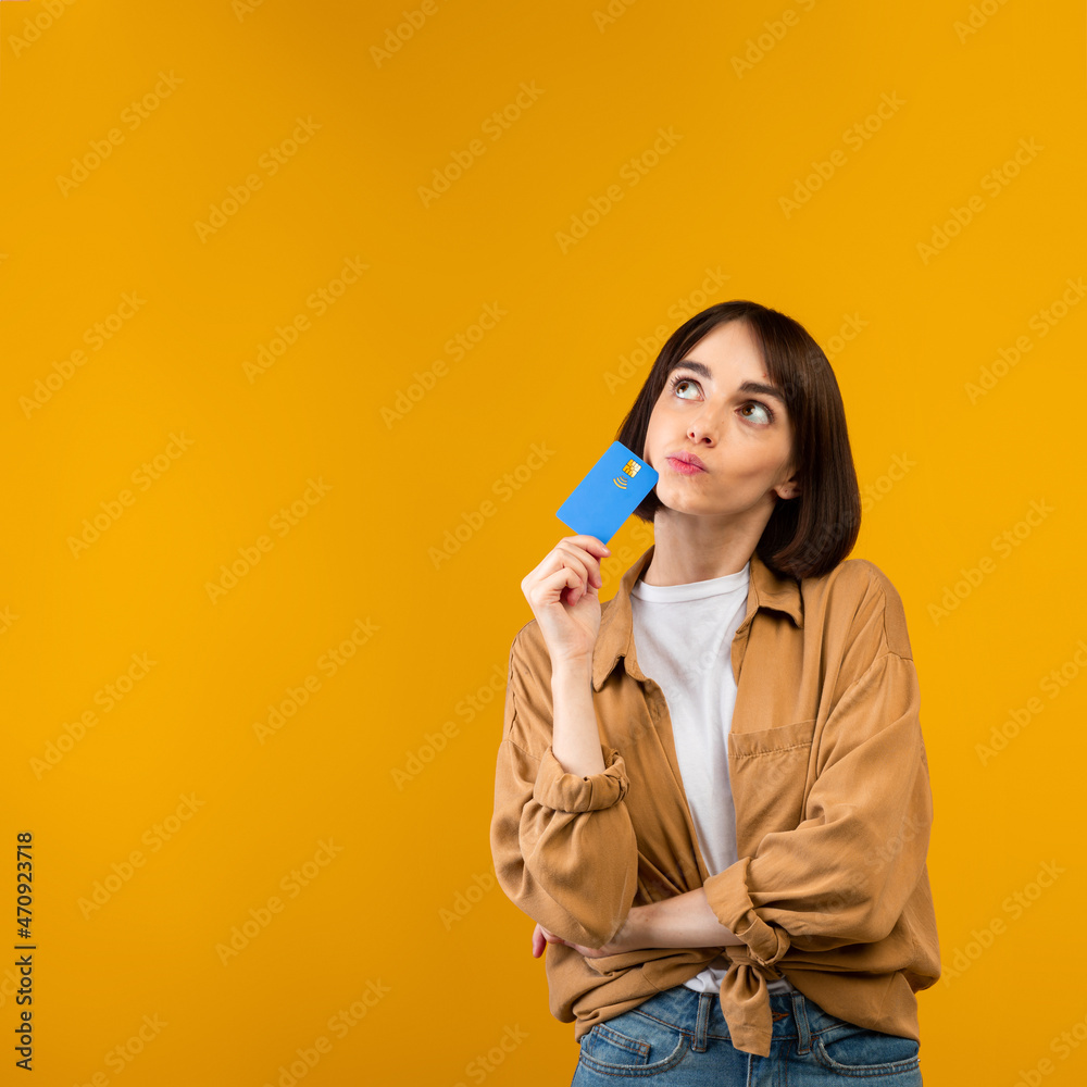 Unlimited shopping concept. Dreamy young woman holding credit card and thinking what to buy, yellow background