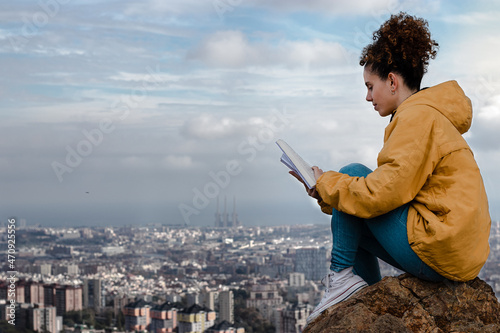 Foto Chica con rizos leyendo libro en montaña con fondo de Barcelona y nubes