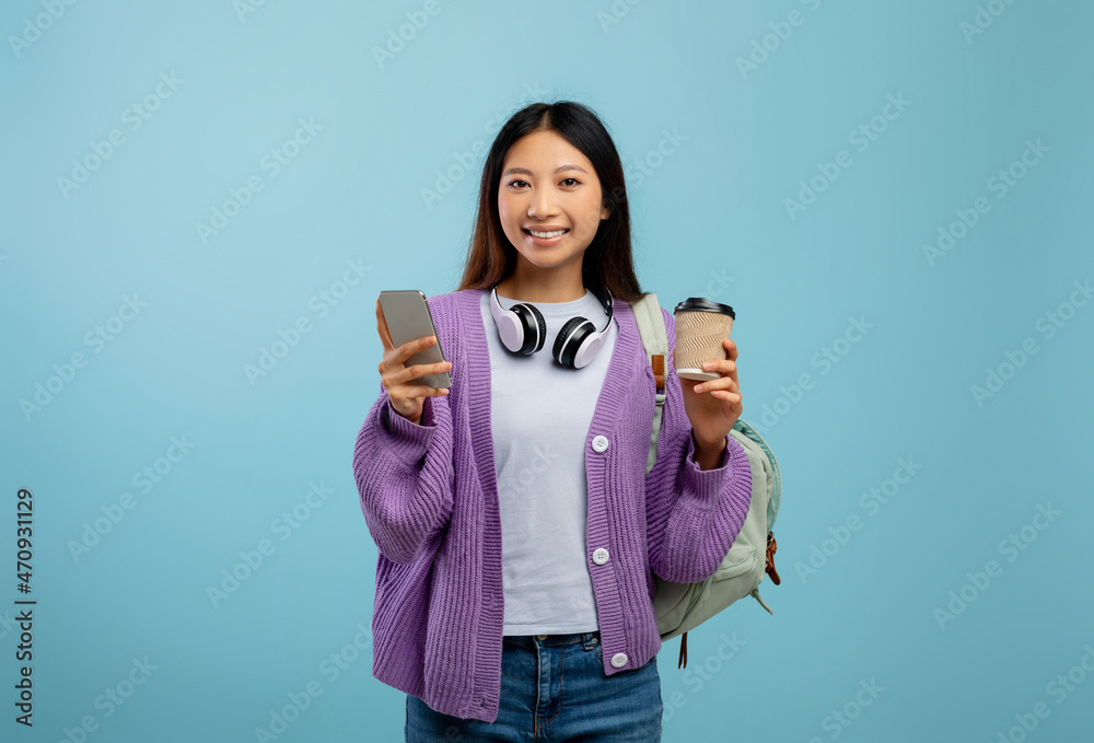 Positive asian lady using smartphone, posing with backpack and holding coffee to go, smiling over blue background Positive asian lady using smartphone, posing with backpack and holding coffee to go, smiling over blue background