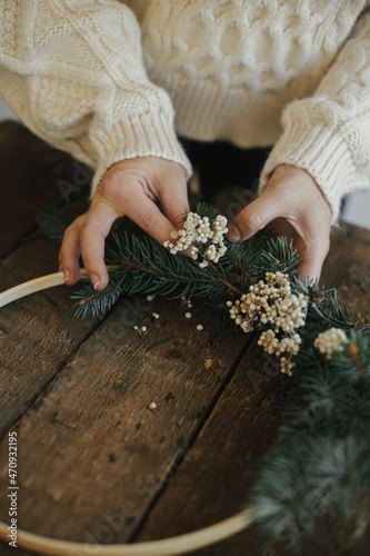 Making modern christmas wreath. Hands in cozy sweater making xmas boho wreath with fir branches, herbs, round wooden hoop on rustic table. Atmospheric moody image. Winter holidays preparation
